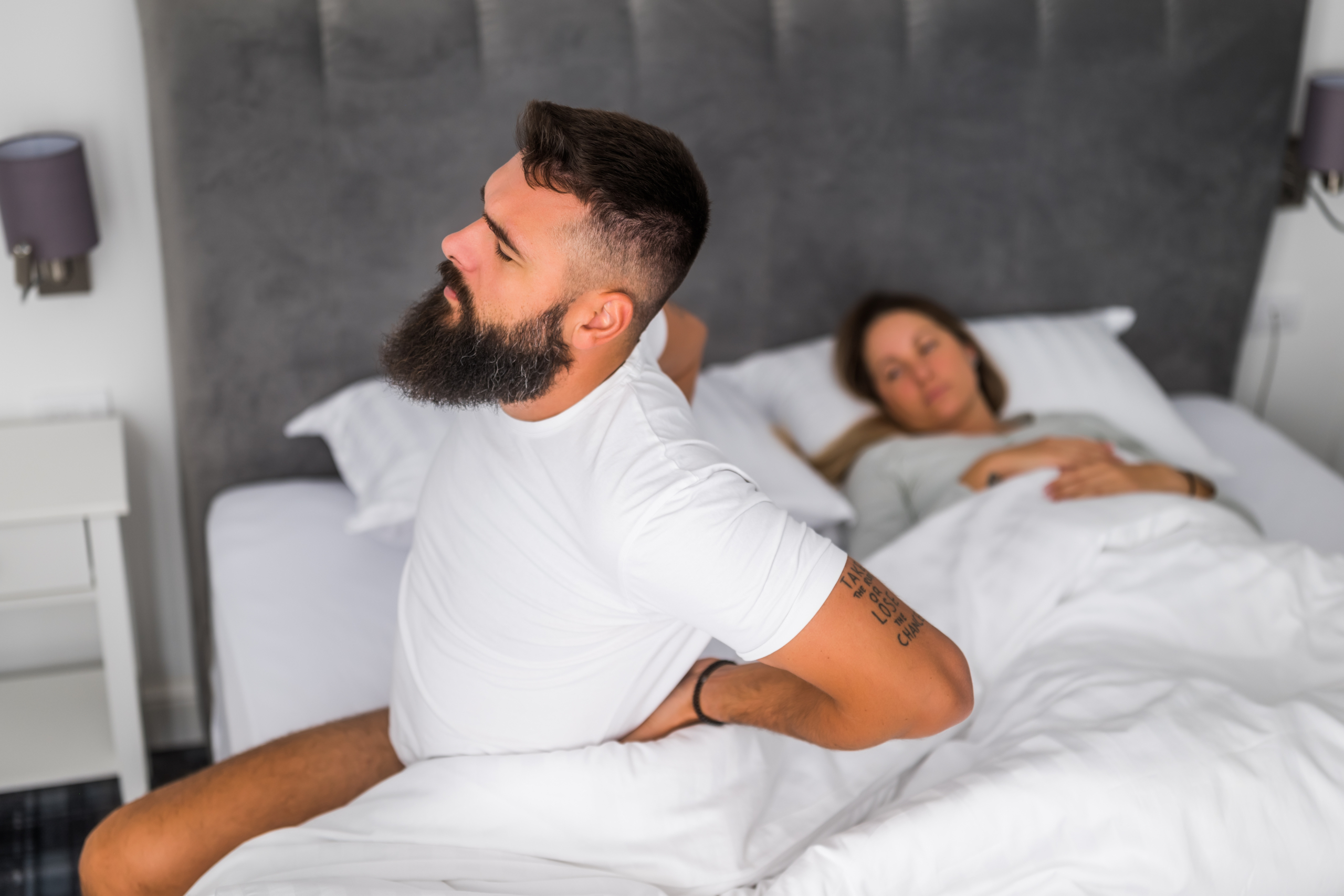 Person experiencing middle back pain while sleeping, sitting on bed and holding their thoracic spine area